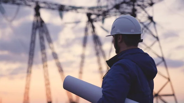 Yüksek elektrik santrallerinin arka planında bir elektrikçi elektrik yapısının geliştirilmesi projesine bakıyor, kablolardaki elektrik voltajının genişletilmesi