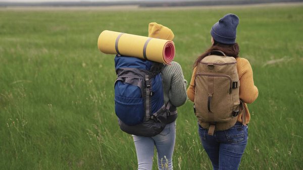 Happy active hipster girls with backpacks on their backs travel through a beautiful valley, a millennial woman on a green field, travel hiking, hiking on the trail, outdoor recreation