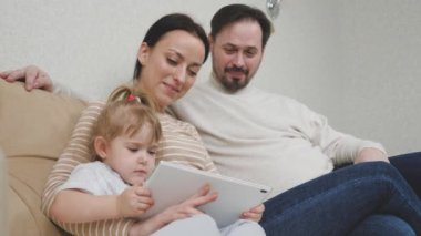 Küçük bir çocuk, annesi ve babasıyla kanepede oturuyor ve tablet oynuyor, çocuk bir alet ekranında eğitim videosu izliyor, bebek için internet uygulamaları, modern aile kavramı.