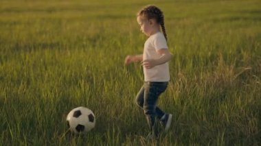 Küçük çocuk gökyüzünde günbatımında futbol oynayarak yeşil futbol sahasında koşuyor. Çocuk topu tekmeliyor ve ona yetişiyor. Mutlu bir çocukluk hayatı, aktif sağlıklı bir bebek.
