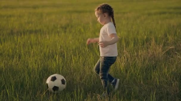 Bambino piccolo corre attraverso il campo di calcio verde giocare a calcio al tramonto nel cielo, bambino calcia la palla e lo raggiunge, il concetto di una vita felice infanzia, bambino sano attivo — Video Stock