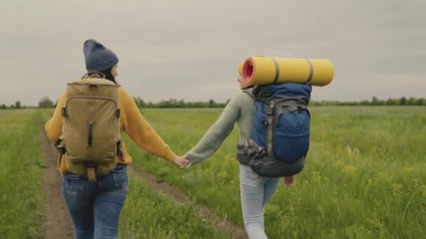 Twee hipster meisjes met rugzakken gaan op een wandeling hand in hand, gelukkige duizendjarige reizigers lopen langs een onverharde weg en op zoek naar avontuur, tijd om te dromen in de natuur, actieve mensen — Stockvideo
