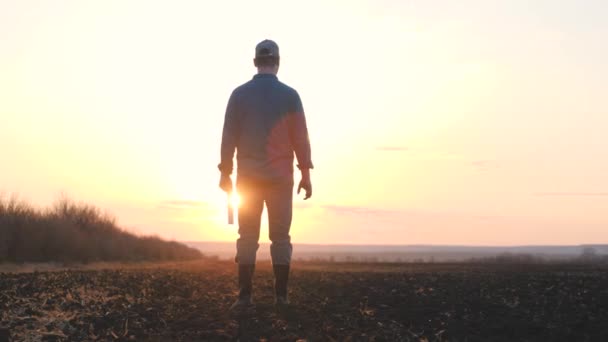 A farmer in boots walks in the sunset on the road with a tablet, the use of modern gadgets in agriculture, work on a land farm, agricultural work in the garden, vegetable garden season — Stock Video