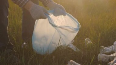 Koruyucu eldivenli bir adam elleri çöp torbasında çöp toplar, doğada gönüllü olarak çalışır, plastik şişeleri, bardakları, yeşil çimlerin üzerindeki peçeteleri temizler, çevre kirliliği