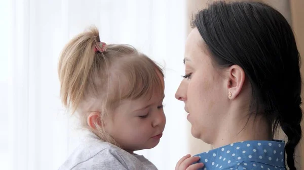 Äiti pitää tytärtään sylissään seistessään ikkunan vieressä ja leikkii nenällään, onnellisella perhe-elämällään, tytöllä äitinsä kanssa viikonloppuisin sisätiloissa, aikuinen lastenhoitaja huolehtii pienestä lapsesta. — kuvapankkivalokuva