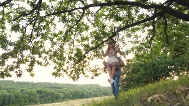 Far rullar sin vuxna dotter på en gunga och skrattar, konceptet med en lycklig familj, pappa och en flicka har kul i naturen, sväva i luften ovanför marken, extrem sommar kul — Stockvideo