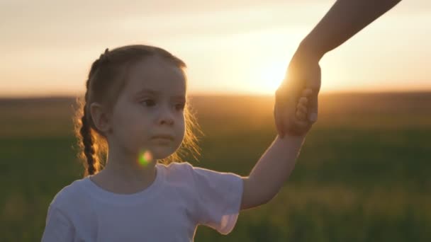 Η μαμά οδηγεί ένα μικρό κορίτσι από το χέρι, κουνώντας το χέρι της στο φως του ηλιοβασιλέματος, ένα όνειρο να είναι μια ευτυχισμένη οικογένεια, ένα κοντινό πλάνο ενός χαμογελαστού παιδιού, κρατώντας ένα παιδί χέρι σε ένα μεγαλύτερο χέρι, μητρική αγάπη και — Αρχείο Βίντεο
