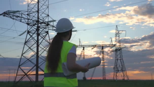 Een elektricien op de achtergrond van hoge torens van elektriciteitscentrales kijkt naar het project voor de ontwikkeling van een elektrische structuur, de uitbreiding van de elektrische spanning van volt in de draden — Stockvideo