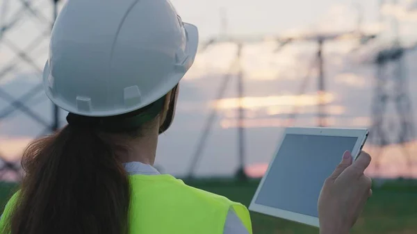 An adult woman electrician is working with a tablet on modern technologies next to a power plant, setting up energized stations through satellite control of networks, volt-installed towers, a gadget