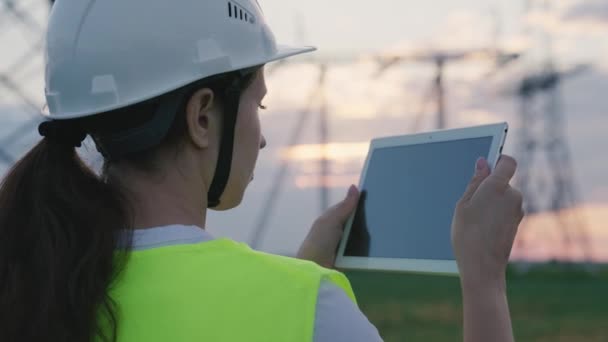 Een volwassen vrouw elektricien werkt met een tablet op moderne technologieën naast een elektriciteitscentrale, het opzetten van energiecentrales door middel van satellietcontrole van netwerken, volt-geïnstalleerde torens, een gadget — Stockvideo