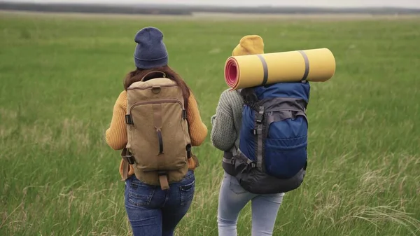 Happy active hipster girls with backpacks on their backs travel through a beautiful valley, a millennial woman on a green field, travel hiking, hiking on the trail, outdoor recreation — Stock Photo, Image