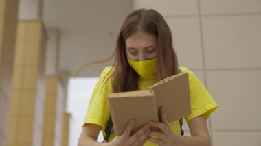 A schoolgirl girl in a mask reads a book while standing at the school, protection against coronavirus, covid-19 pandemic, air filtration through a mask filter, modern education for adolescents clipart
