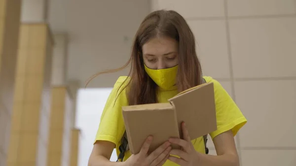 Maskeli bir kız, okulda dururken kitap okuyor. Koronavirüse karşı koruma, covid-19 salgını maske filtresinden hava filtresi, ergenler için modern eğitim.