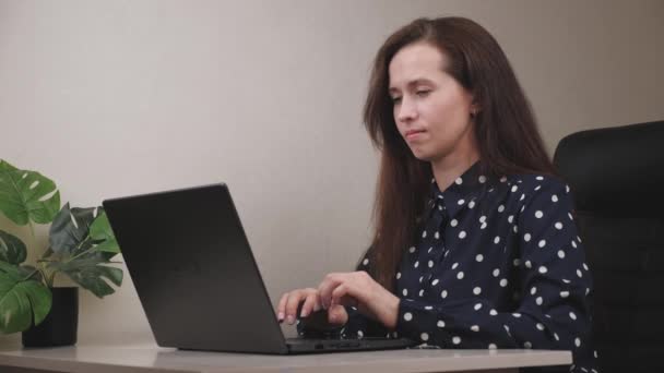 Une fille gentille à un ordinateur portable tapant du texte dans un ordinateur portable, souriant et prêtant main forte, un employé de bureau, une femme d'affaires organise un lieu de travail à un ordinateur, travail indépendant dans un bureau à domicile — Video