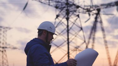 Yüksek elektrik santrallerinin arka planında bir elektrikçi elektrik yapısının geliştirilmesi projesine bakıyor, kablolardaki elektrik voltajının genişletilmesi