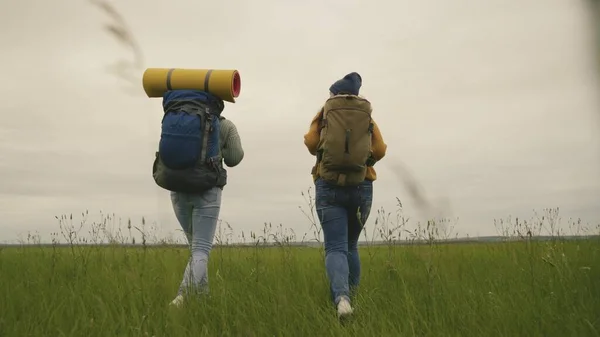 Chicas hipster activas felices con mochilas en la espalda viajan a través de un hermoso valle, una mujer milenaria en un campo verde, excursiones, senderismo en el sendero, recreación al aire libre —  Fotos de Stock