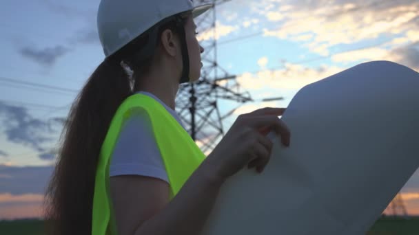 Eine Frau Power Engineering mit einem Projekt der neuen elektrischen Werke, eine Elektrikerin geht vor dem Hintergrund der hohen Türme von Kraftwerken, die Ausweitung der elektrischen Ausrüstung von Spannung Volt — Stockvideo