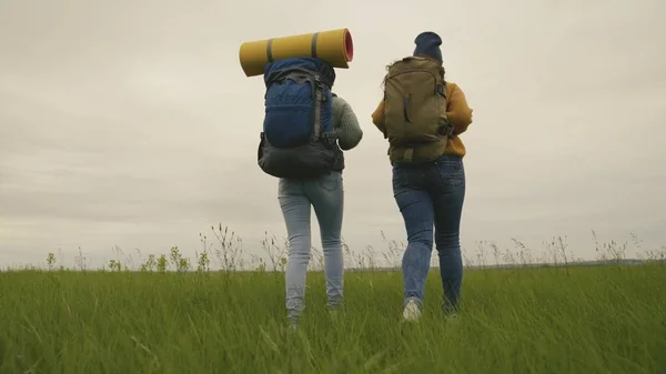 Chicas hipster activas felices con mochilas en la espalda viajan a través de un hermoso valle, una mujer milenaria en un campo verde, excursiones, senderismo en el sendero, recreación al aire libre —  Fotos de Stock