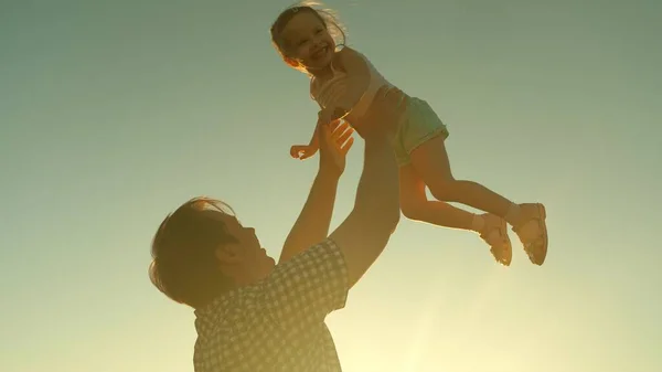 Tata wyrzuca szczęśliwe dziecko w niebo o zachodzie słońca. Córka taty lata w jego ramionach wznosząc się w powietrzu grając w samolot gry w blasku słońca. Dzieciak raduje się ze swoim ojcem. — Zdjęcie stockowe