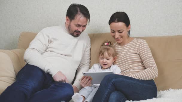 Een klein kind zit op de bank met zijn moeder en vader en speelt een tablet, kind kijkt naar een trainingsvideo in een gadget display, internet toepassingen voor baby, het concept van moderne familie — Stockvideo