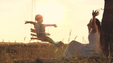 Anne, gün batımında küçük bir çocuğu salıncakta sallıyor, mutlu bir aile, sevgi ve bir çocuğa bakıyor, kolları açık havada süzülüyor, çocukluk hayali uçuyor, güneş ışığında oyun oynuyor.