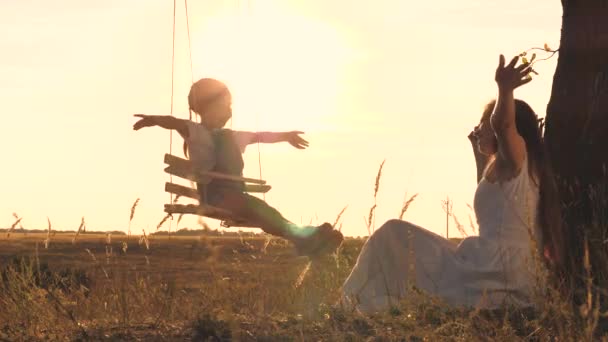 Mamá enrolla a un niño pequeño en un columpio al atardecer, una familia feliz, el amor y el cuidado de un niño, se elevan en el aire con los brazos extendidos, un sueño de la infancia de volar, juegos en el patio de recreo en el sol — Vídeos de Stock