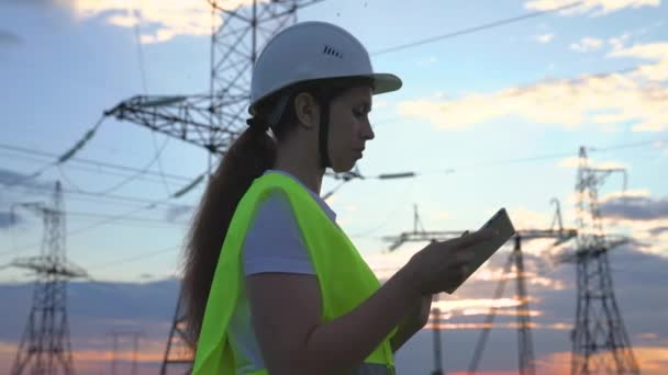 En voksen kvinde elektriker arbejder med en tablet på moderne teknologier ved siden af et kraftværk, oprettelse af energi stationer via satellit kontrol af netværk, volt-installerede tårne, en gadget – Stock-video