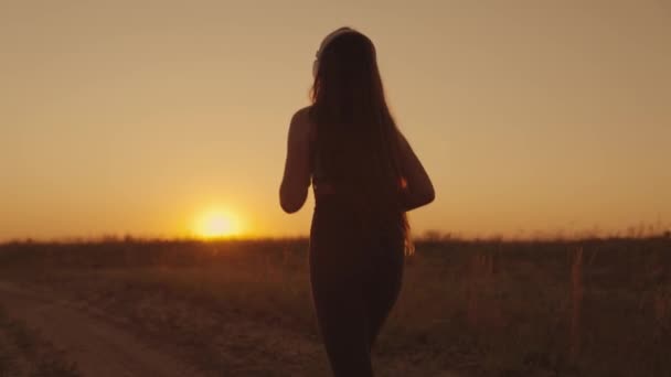 Девушка в наушниках бежит по дороге на закате, молодая женщина делает утреннюю пробежку, здоровый образ жизни на свежем воздухе, междугороднюю гонку, подготовку к победе в спортивных соревнованиях — стоковое видео