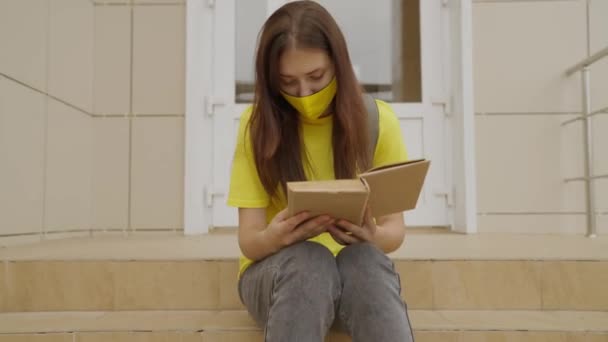 A schoolgirl girl in a mask reads a book while standing at the school, protection against coronavirus, covid-19 pandemic, air filtration through a mask filter, modern education for adolescents — Stock Video