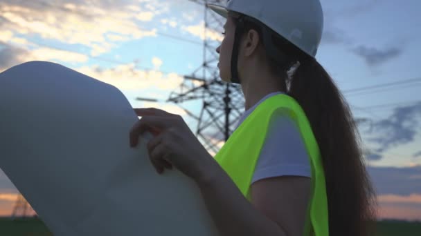 Una mujer ingeniero de energía con un proyecto de nuevas obras eléctricas, un electricista camina sobre el fondo de torres altas de plantas de energía, la expansión de equipos eléctricos de voltios de voltaje — Vídeos de Stock
