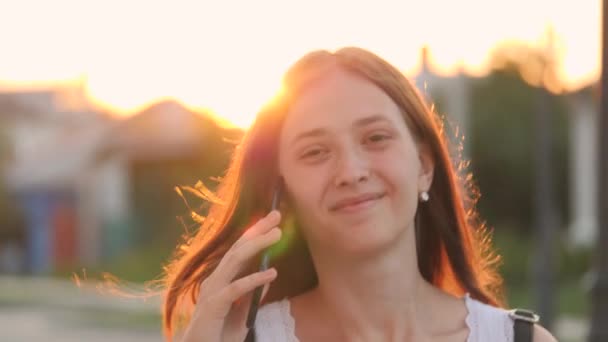 Chica feliz caminando por las calles de la ciudad y hablando en el teléfono inteligente en el resplandor de la luz del sol, estudiante universitario alegre en el resplandor de la puesta del sol, concepto de negocio, tecnología de teléfono táctil — Vídeos de Stock
