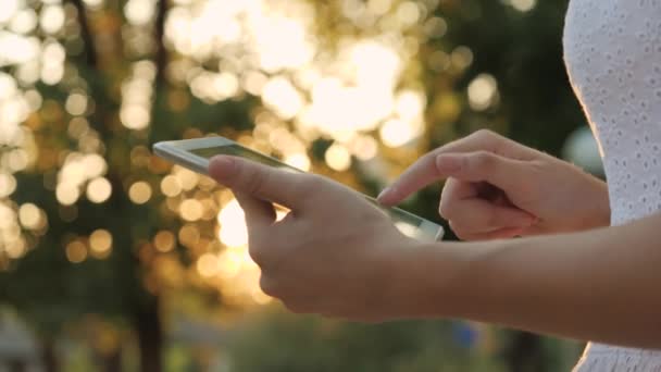 Una giovane ragazza tiene in mano un tablet moderno nell'abbagliamento del tramonto, va online su Internet, i raggi del sole brillano sul touch screen del tablet, il concetto di tecnologie moderne — Video Stock