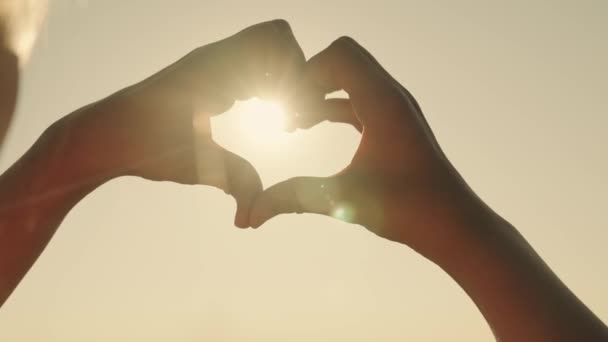 Mostrar com gestos de mão coração no fundo do céu no brilho do pôr do sol, o sonho de amor e saúde, um raio de sol, uma menina bonita no brilho da luz de verão, conceito — Vídeo de Stock