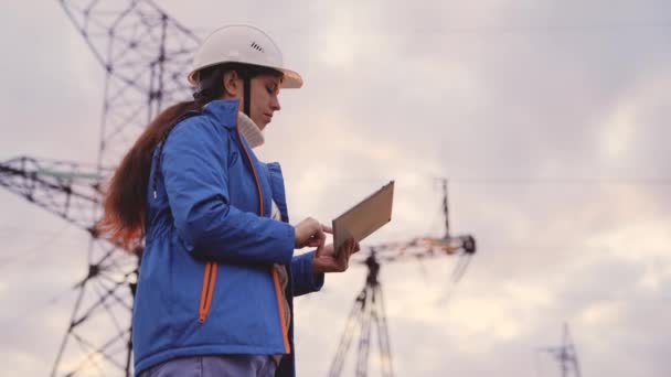 An electrician engineer in helmet works with tablet at sunset in sky, tower under high voltage current, checking electrical volt networks, supplying light to city, industrial safety power engineer — Stock Video