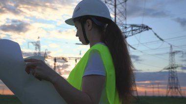 Yeni elektrik tesisleri projesine sahip bir kadın elektrik mühendisi, bir elektrikçi, yüksek elektrik santrallerinin arka planında yürür, elektrik gereçlerinin genişletilmesi,