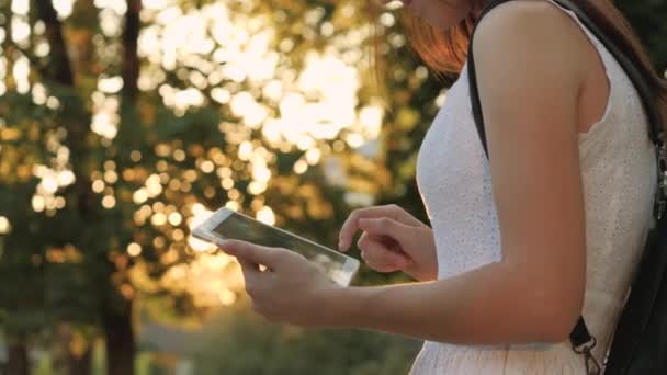 Una joven sostiene en su mano una tableta moderna en el resplandor de la puesta del sol, ir en línea a Internet, los rayos del sol brillan en la pantalla táctil de la tableta, el concepto de las tecnologías modernas — Vídeo de stock