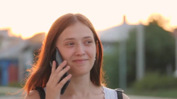 Щаслива дівчина гуляє по міських вулицях і розмовляє по смартфону в відблисках сонячного світла, весела студентка університету в відблисках заходу сонця, бізнес-концепції, технології сенсорного телефону — стокове відео