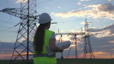 Yüksek kulelerden oluşan bir elektrik santralinde elektrik mühendisi gün batımında elektrik ağlarının performansını kontrol eder. Güçlü yüksek voltajlar, enerji alanında çalışan bir adam..