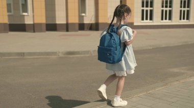 Sırt çantalı küçük bir kız okul bahçesinde okula gidiyor, ilkokul çocukları için eğitim programı kaldırımda yürüyor ve eğleniyor, çocukluk hayalleri kurarak yeni bir hayat kuruyor.