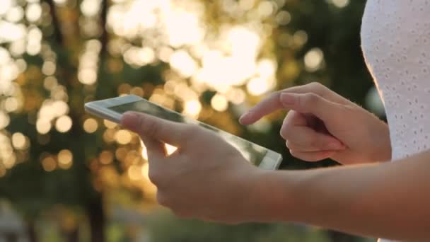 Ein junges Mädchen hält im gleißenden Sonnenuntergang ein modernes Tablet in der Hand, geht ins Internet, die Sonnenstrahlen scheinen auf den Touchscreen des Tablets, das Konzept der modernen Technologien — Stockvideo