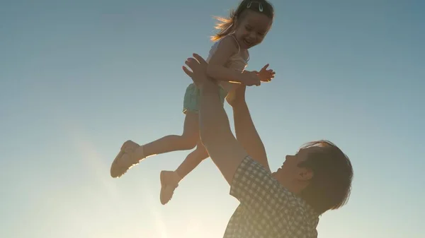 Szczęśliwy ojciec rzuca dziecko w niebo z blaskiem słońca, dziecko leci wysoko jak samolot i śmieje się, dziewczynka bawi się na świeżym powietrzu ze spapą, koncepcja szczęśliwej rodziny — Zdjęcie stockowe