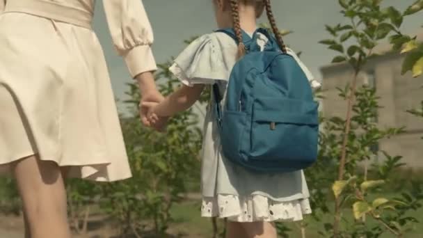 Madre lleva a un niño pequeño de la mano al primer grado de la escuela primaria, bolsa de la escuela en los hombros de una niña, familia feliz, apresurarse a la lección en el patio de la escuela, niño con mochila recibirá una educación — Vídeos de Stock