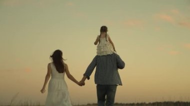 Gün batımında küçük bir çocukla mutlu bir aile, tüm aileyle bir tatil gezisi, baba ve çocuk neşeyle oynuyorlar, acele ediyorlar, anne ve baba çocuklarını omuzlarında taşıyorlar.