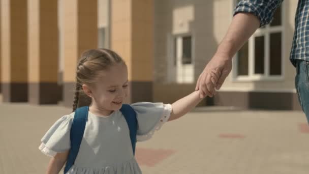 Papà conduce il bambino a mano a scuola, primo piano, una piccola scolaretta con lo zaino, una famiglia felice, un sogno d'infanzia dei bambini, una studentessa attiva, una buona educazione moderna, ama e ricevi la conoscenza — Video Stock