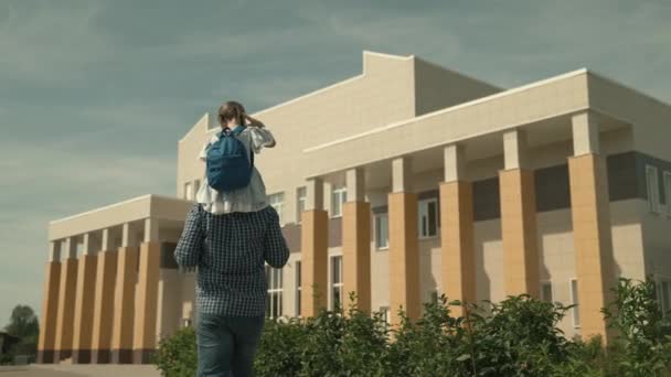 Een vader draagt een dochtertje op zijn schouders naar school, een meisje gaat naar school, gelukkig gezin, zorgzame vader leidt het kind naar een onderwijsinstelling voor kinderen, om een opleiding te krijgen, kind eerste klas — Stockvideo