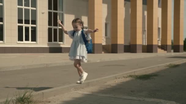 Glückliches kleines Mädchen, das auf dem Schulhof spielt, ein kleines Kind mit einem Rucksack, einer Kinderschultasche auf den Schultern der Babys, einer lustigen Kindergrundschulbildung, einem Kindheitstraum, einem lustigen Schulleben — Stockvideo