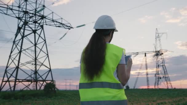 An electrician works with a tablet at a large power plant supplying electricity, an energy engineer makes a project of high voltage towers, run current through wires using modern technologies — Stock Video