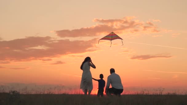 Família feliz, criança pequena com o pai e a mãe lançam o papagaio voador no céu do por do sol, silhueta da mãe pai e da criança junto, moscas de brinquedo voando altamente em sua cabeça, sonhos da fantasia das crianças, divertimento da viagem — Vídeo de Stock