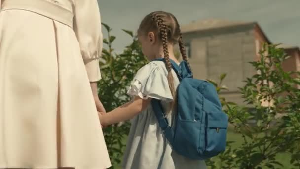 Mamá lleva a su hija pequeña con una mochila a la escuela, escolta al niño a la educación preescolar, camina a casa a través del patio de la escuela, estudiante de primer grado, familia feliz, niño con el bolso de la escuela a mano con la madre — Vídeos de Stock