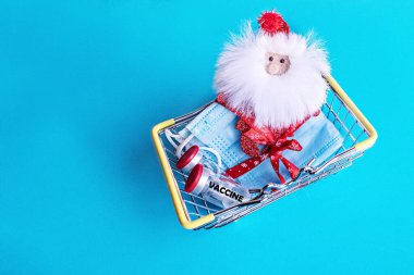 Minyatür bir alışveriş sepetinde tıbbi koruyucu maskeler, şenlikli bir kurdele ve mavi arka planda koronavirüs aşısı olan Noel Baba ile bağlanmış. Yeni Yıl ve Noel konsepti.